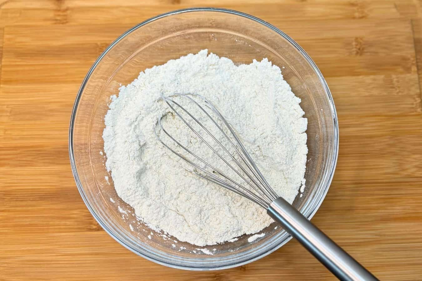 A glass mixing bowl filled with dry pancake ingredients and a metal whisk on top, sitting on a wooden surface.