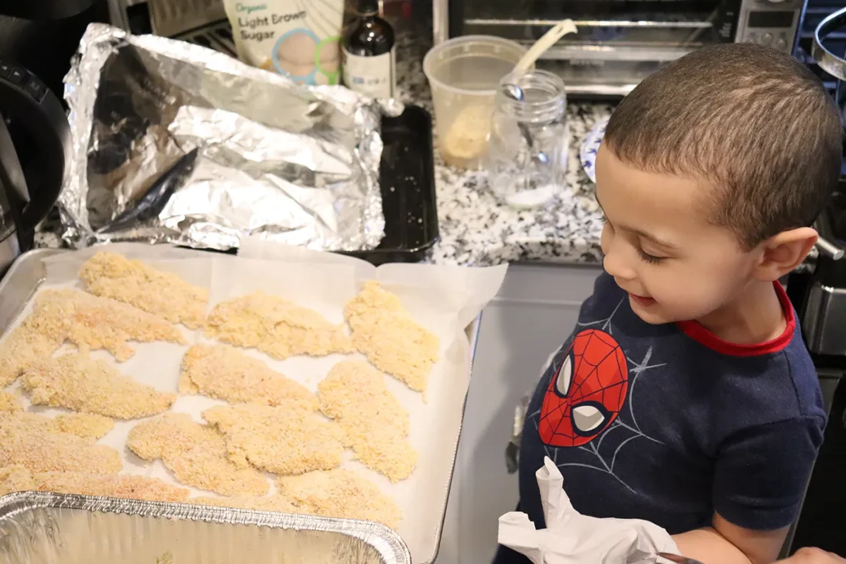 The Benefits of Cooking With Kids: Young child helping in the kitchen by preparing breaded chicken on a baking tray.