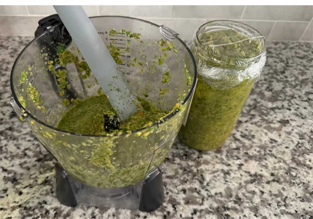 A food processor filled with sazón next to a jar covered with plastic wrap, storing the green seasoning.