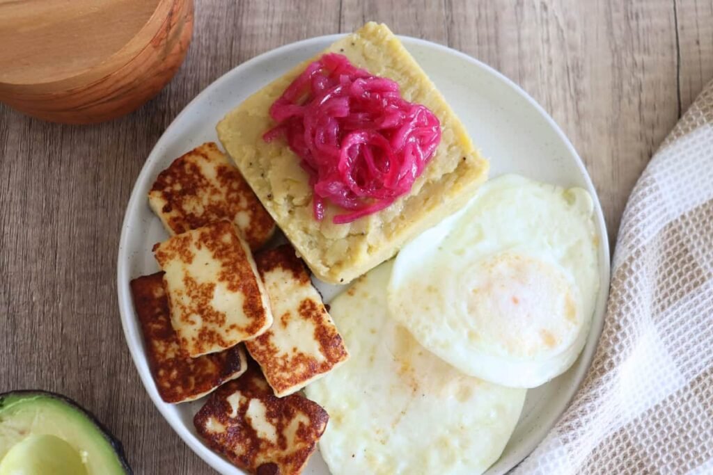 A Dominican breakfast plate with mashed mangú topped with pickled red onions, fried cheese, and sunny-side-up eggs.