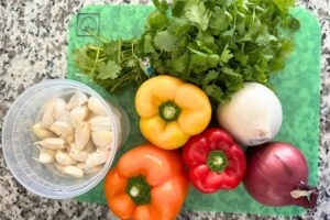 Fresh ingredients for Dominican sofrito (sazón) laid out on a cutting board, including garlic, bell peppers, onions, and cilantro.