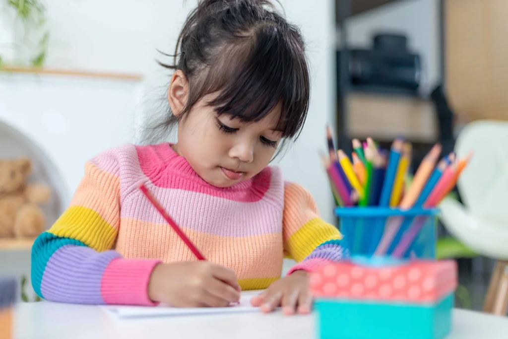 Young girl drawing at a homework station with colored pencils in a rainbow sweater