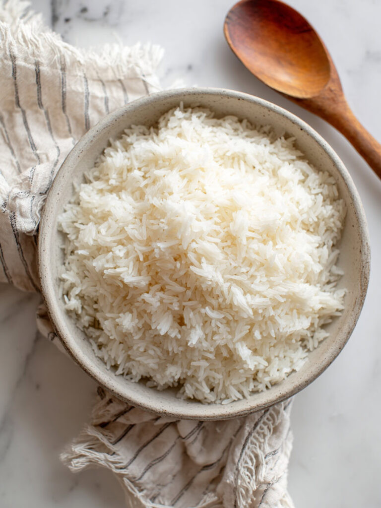 A bowl of fluffy white rice in a rustic ceramic bowl, with wooden spoons and a striped towel beside it on a wooden surface.