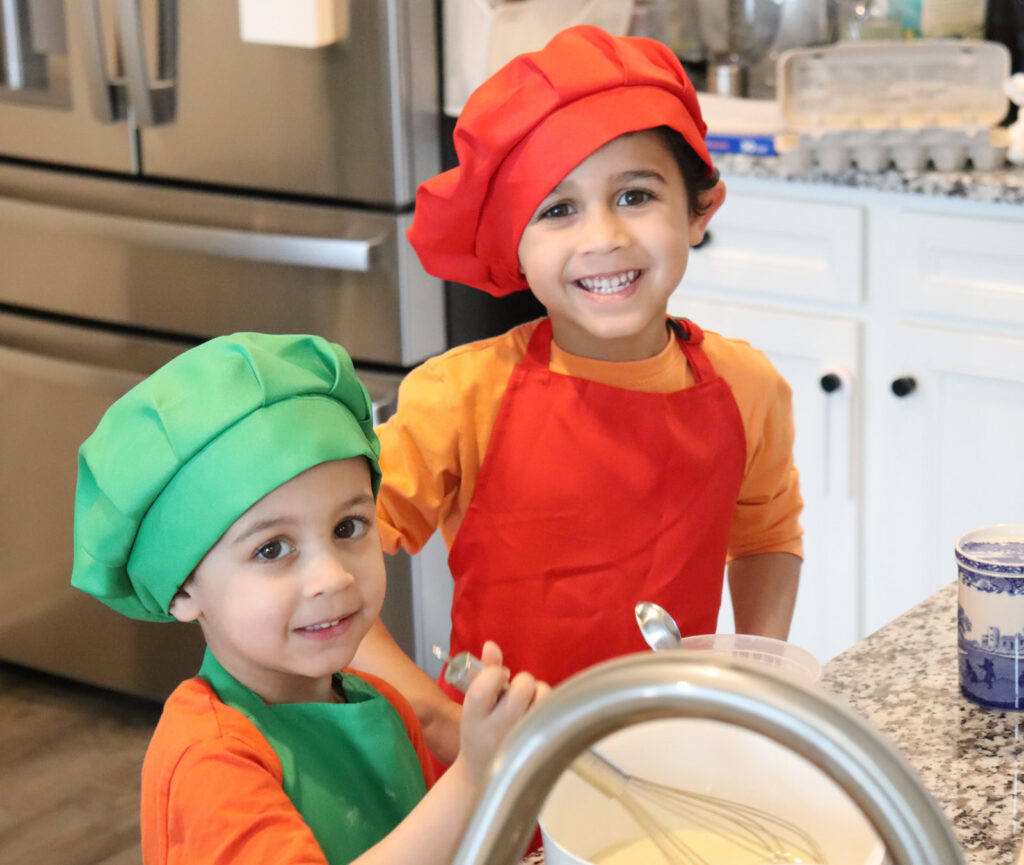 The Benefits of Cooking With your Kids: Two children in chef hats whisking batter in the kitchen, showing why you should involve kids in cooking.