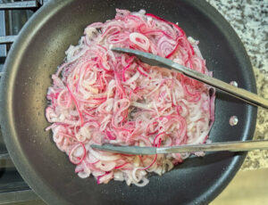 Dominican sautéed onions: onions sautéing in a skillet, stirred with tongs until soft but still with a bite.