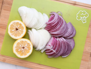 Dominican sautéed onions prep, thinly sliced red and white onions with halved sour orange on a cutting board.