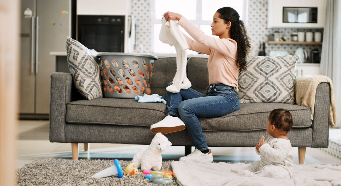 Mother folding laundry on a couch while her baby plays on the floor, representing busy parents using Habit Hooks to manage household routines more efficiently.