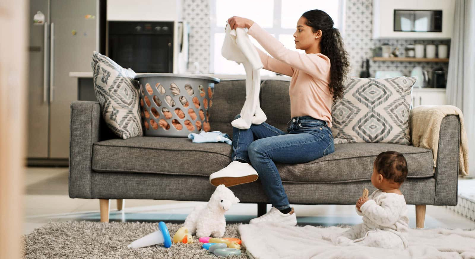 Mother folding laundry on a couch while her baby plays on the floor, representing busy parents using Habit Hooks to manage household routines more efficiently.