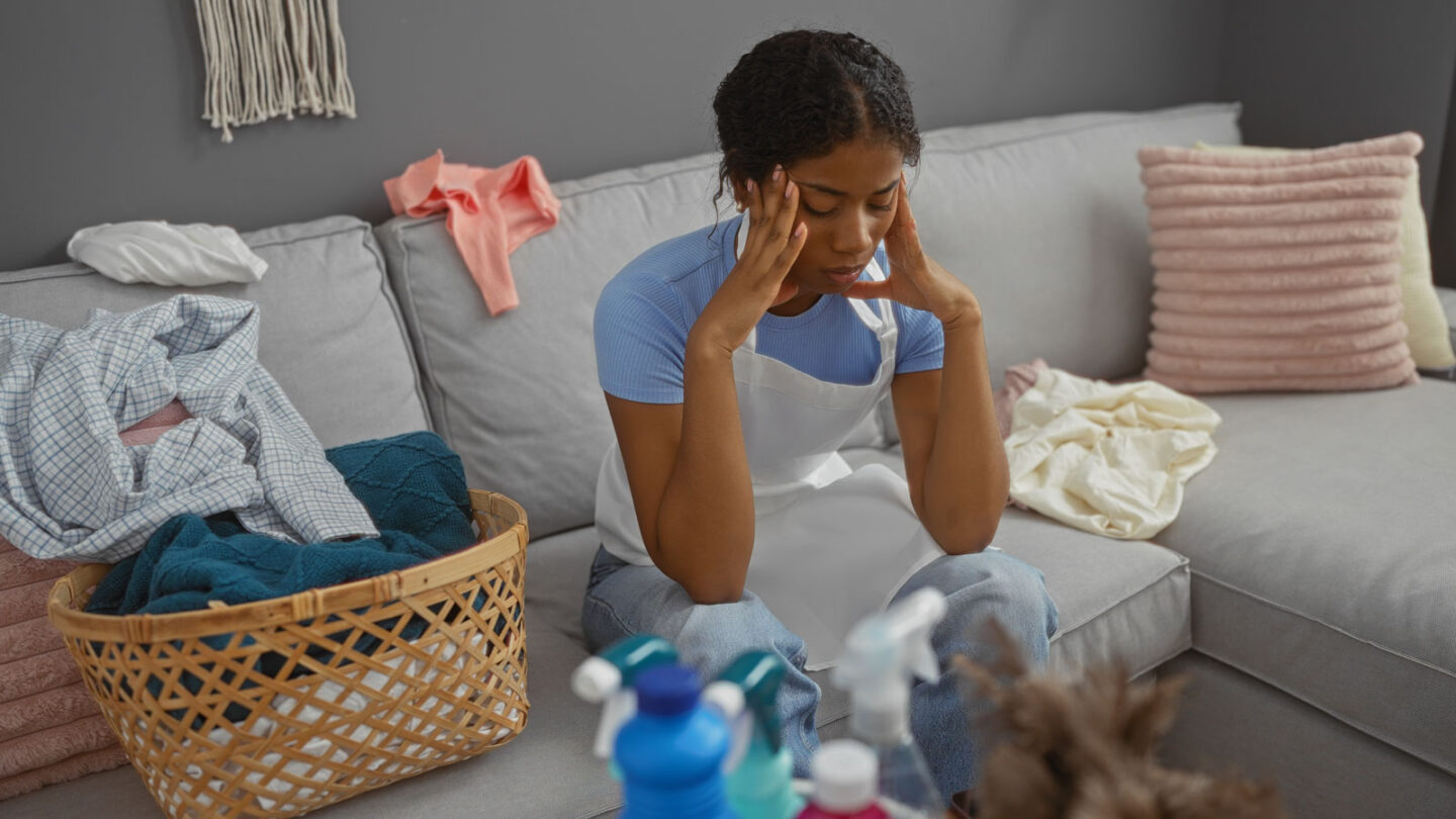 Tired woman sitting on a gray couch surrounded by laundry and cleaning supplies, holding her head in her hands from exhaustion.