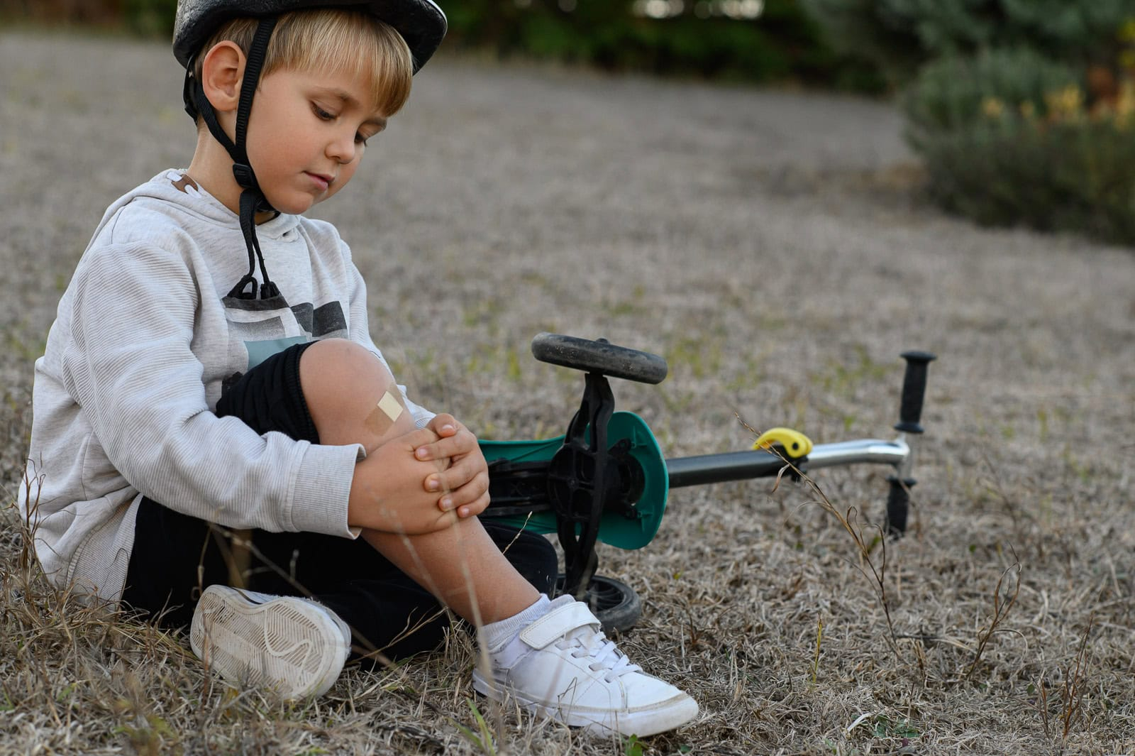 Child wearing a helmet sits on the ground holding a bandaged knee beside a fallen scooter, reflecting resilience after a setback
