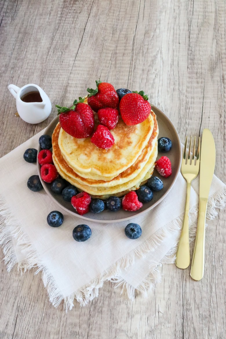 Hero image of fluffy pancakes topped with berries on a plate with syrup pitcher and gold utensils