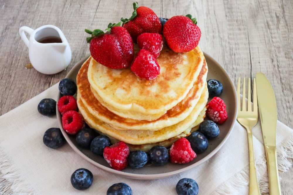 Stack of fluffy pancakes topped with strawberries, raspberries, and blueberries on a plate with syrup