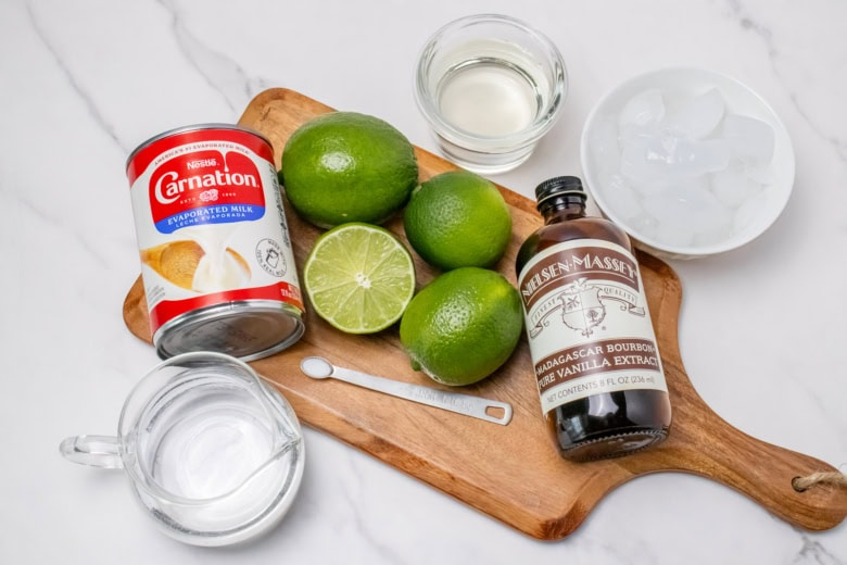 Ingredients for Morir Soñando de Limón including evaporated milk, fresh limes, vanilla, water, and ice on a wooden board.