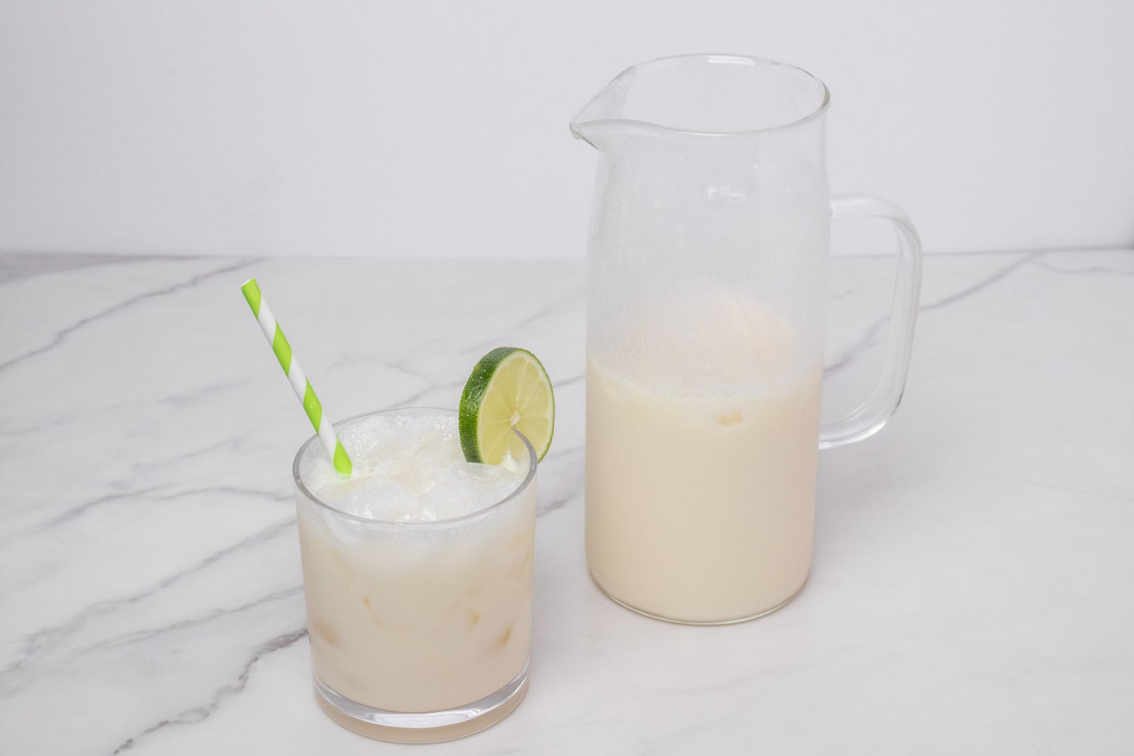Glass of Morir Soñando de Limón over ice with lime garnish and pitcher in the background.
